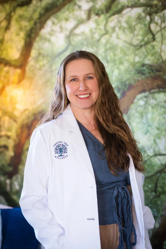Dr Tracy Parrish in a Twisted Tree Wellness white coat, smiling in front of a tree mural