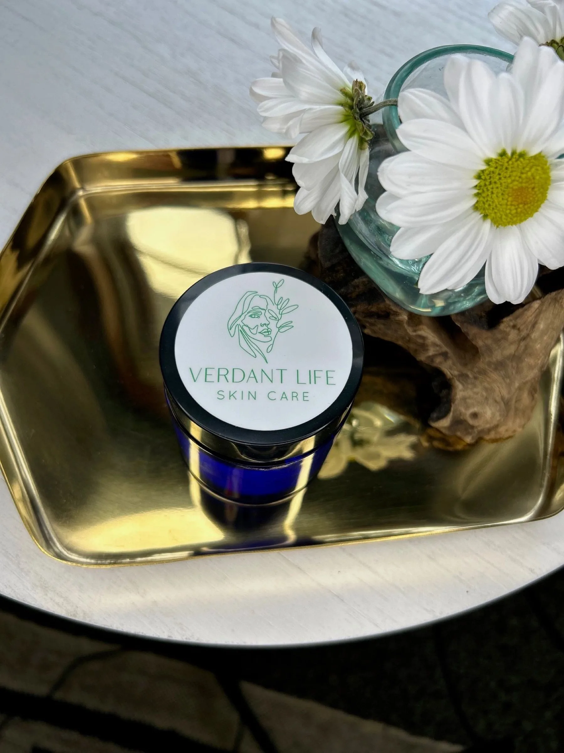 Blue jar of Verdant Life Salt Scrub on a gold tray beside white daisies.