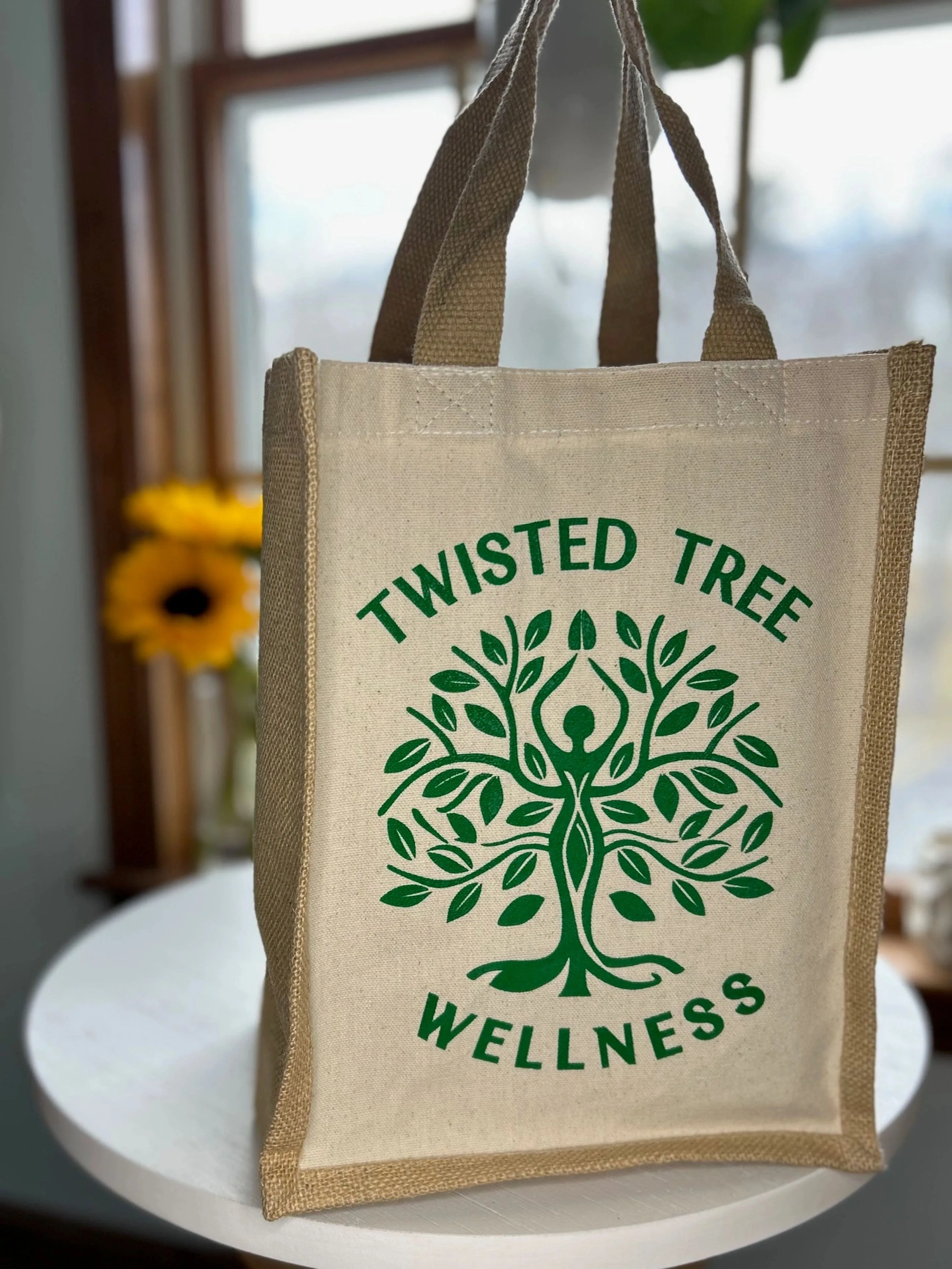 Twisted Tree Wellness jute and canvas tote bag with the green tree logo, on a table by a window