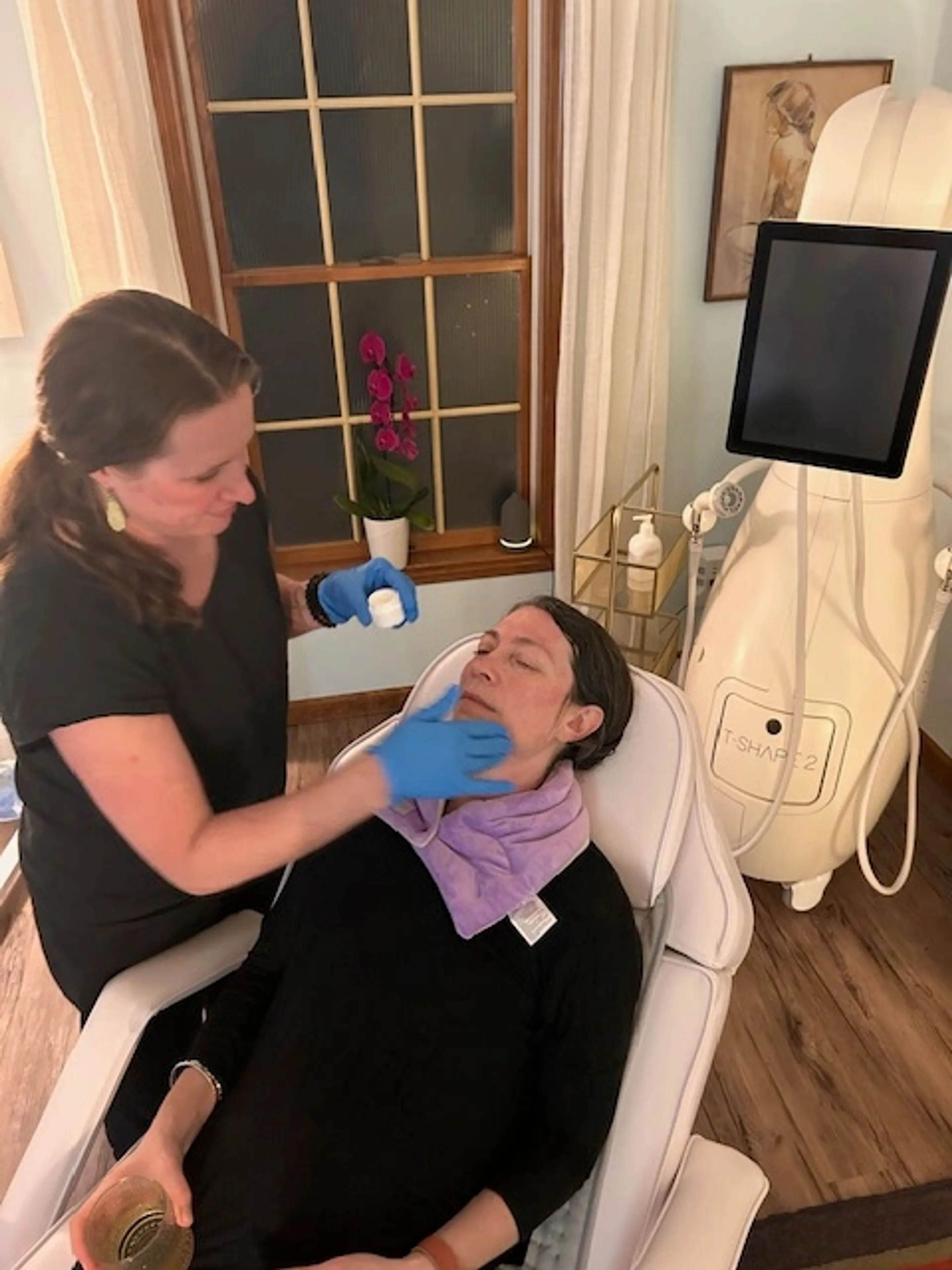 Dr Parrish in blue gloves applying serum to a reclining client's cheek next to the Tshape 2 machine