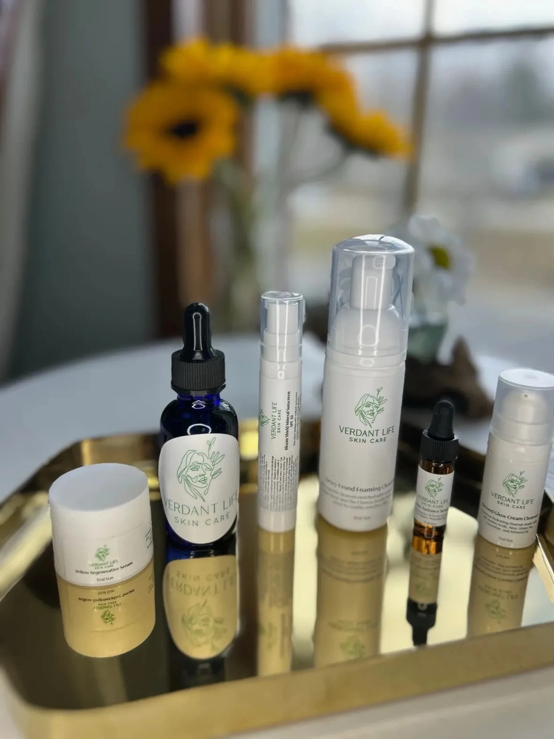 Verdant Life Skin Care bottles and jars on a gold mirrored tray, with sunflowers behind.