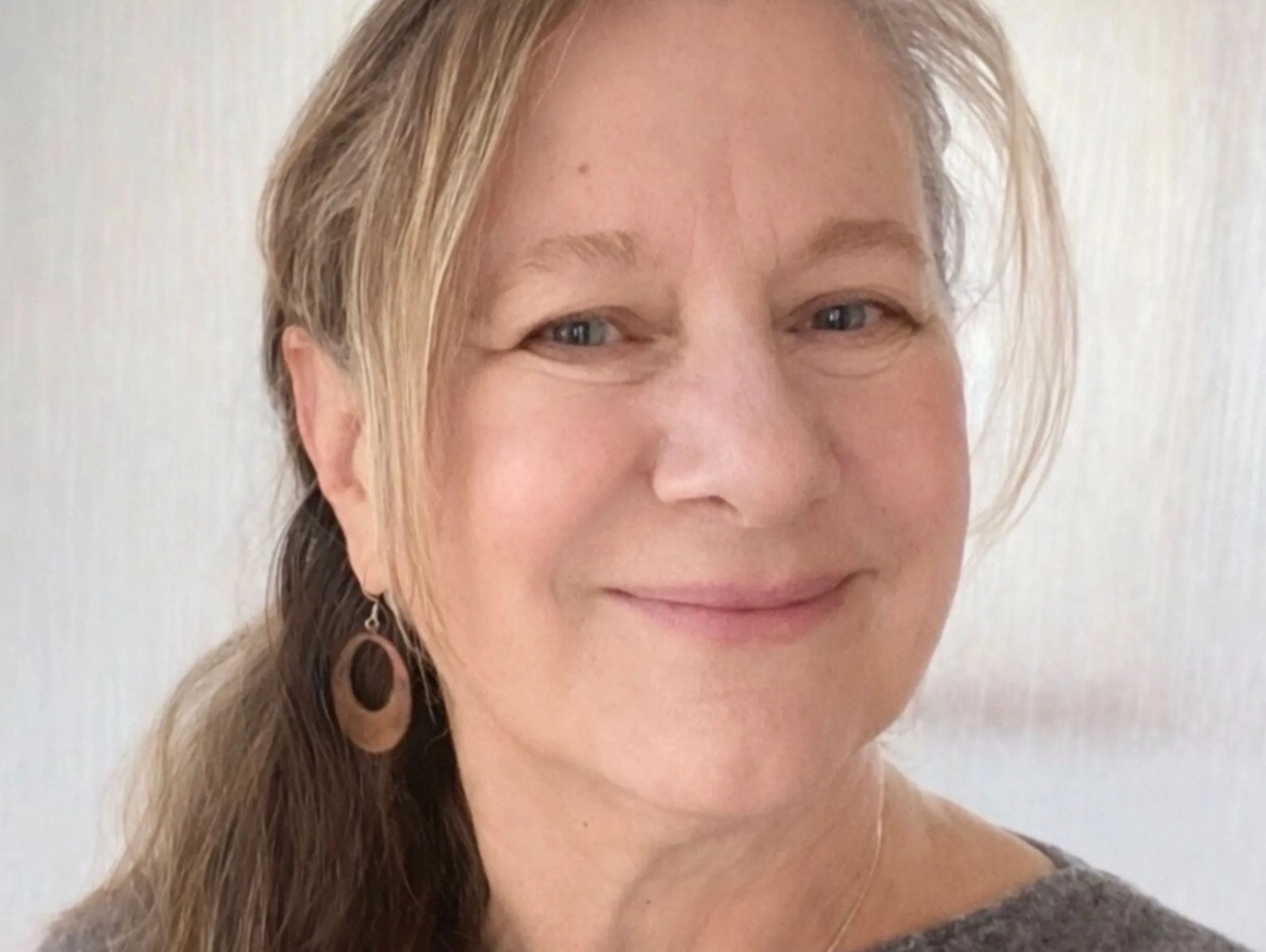 Smiling woman with light brown hair wearing wooden earrings and a gray sweater.